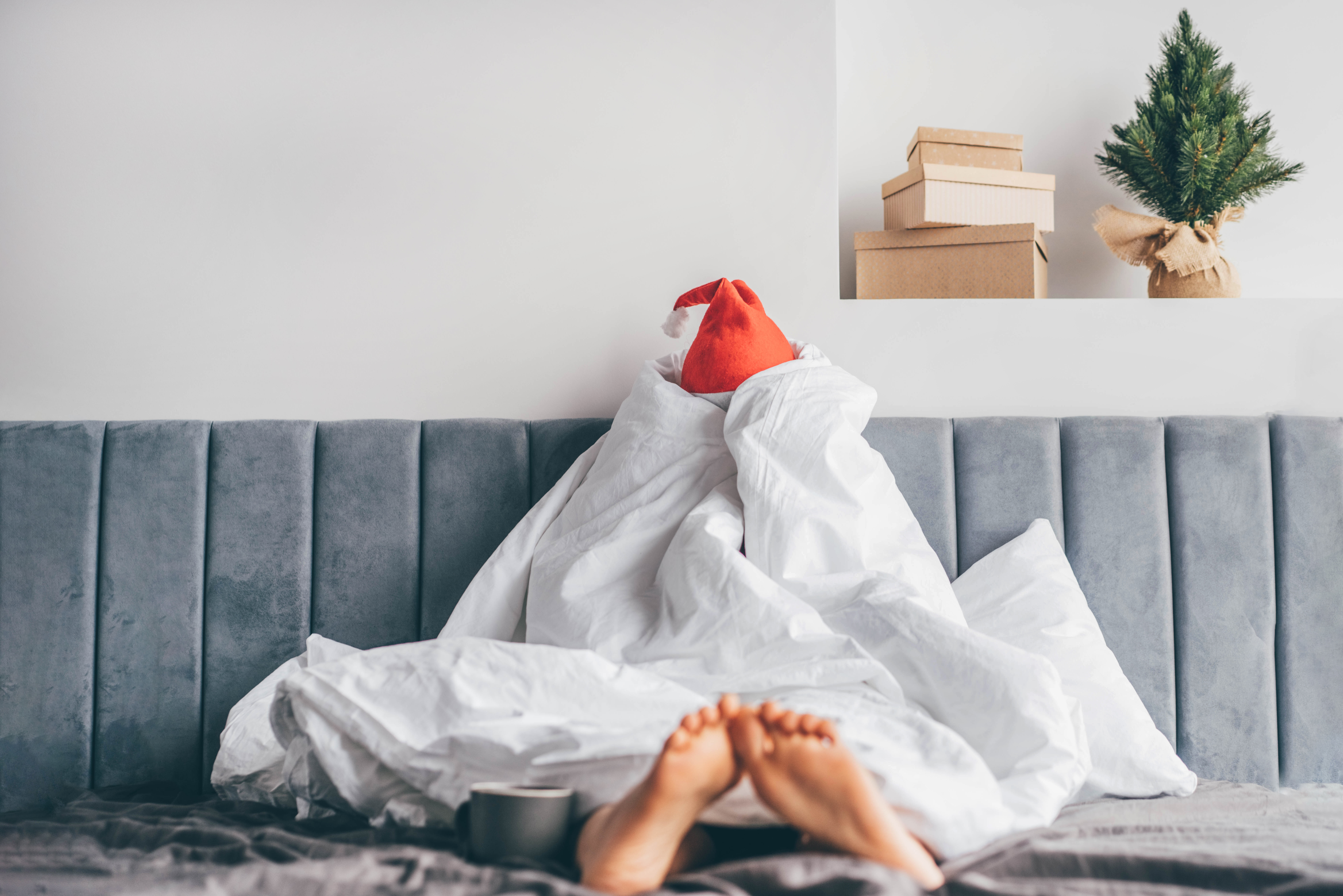 A woman is sitting up in bed, wrapped completely in a white duvet and wearing a Santa hat with a moody, bored expression.
