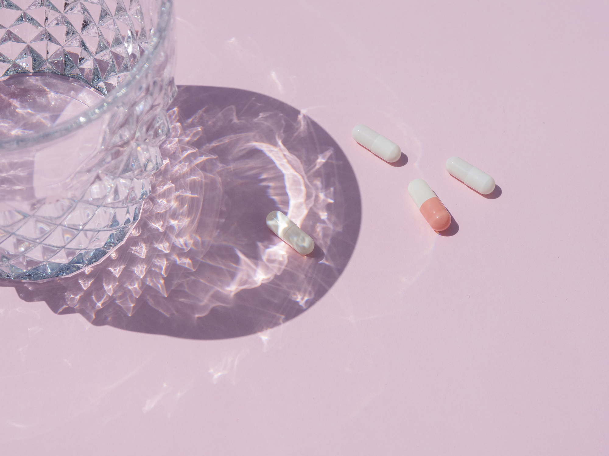 Suplements pills on a pink background next to a glass of water with reflections and shades of sunlight